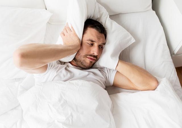 person laying in white bed using a pillow to cover their ears